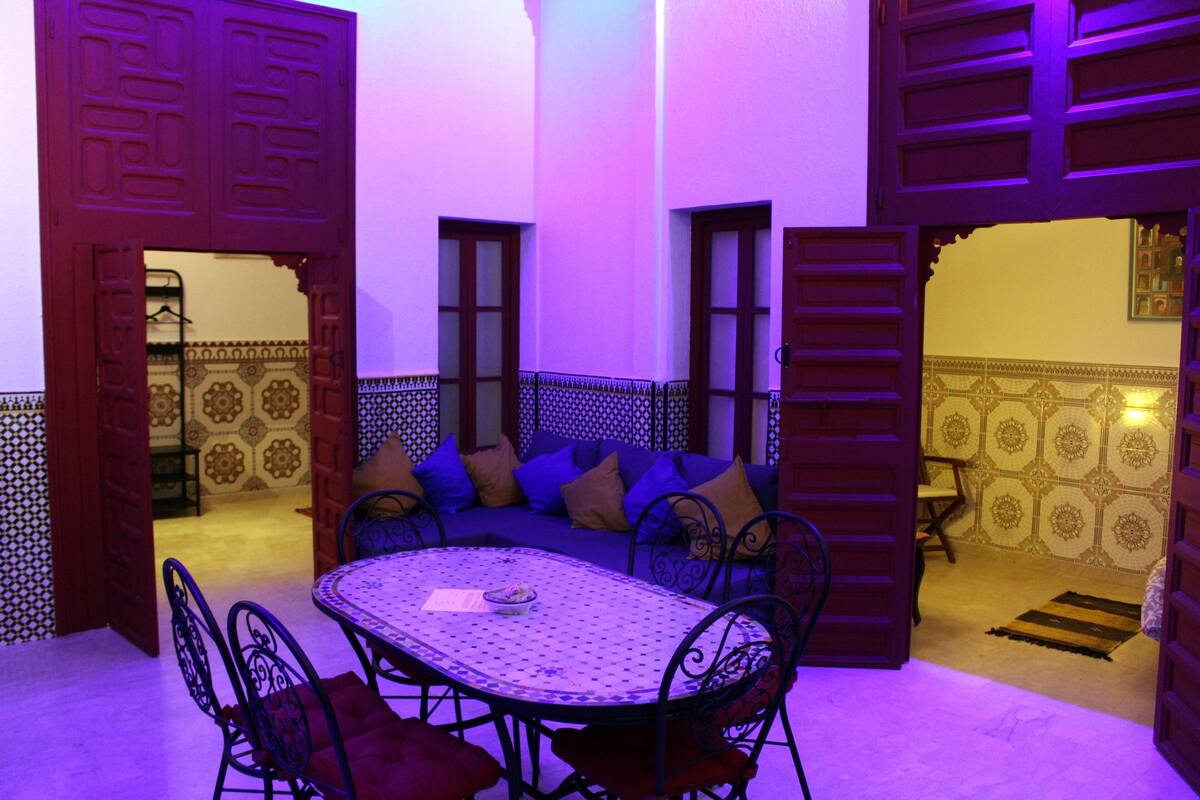 An open-plan living area with a round table and chairs, a sofa with cushions, and patterned walls.