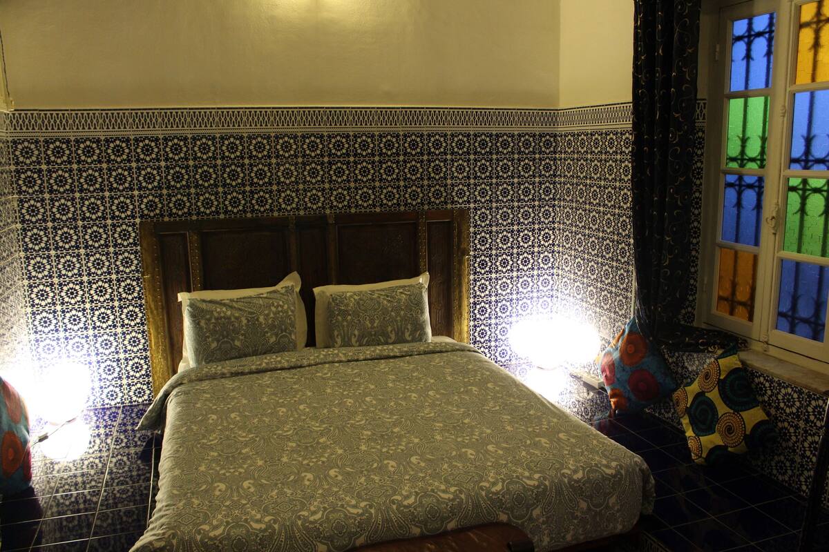 A bed with a patterned bedspread, a headboard, and a window with colored glass panes.