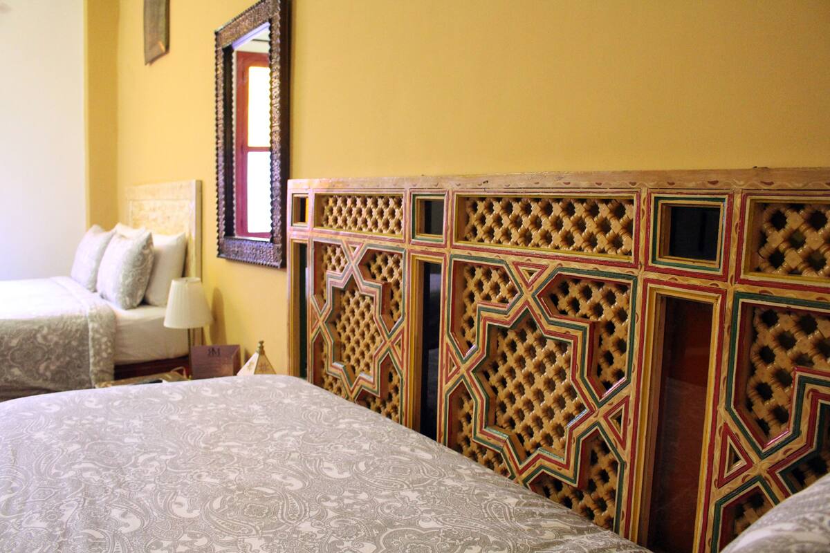 A hotel room with two beds, a patterned headboard, a mirror, and a bedside table with a lamp.