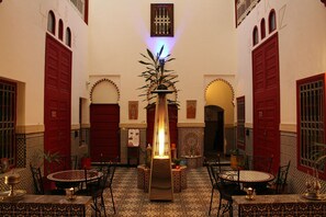 Un intérieur marocain traditionnel avec une fontaine centrale, des carreaux décorés et des portes rouges.
