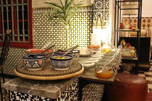 A dining area with a patterned tile wall, a table with bowls and a jar of orange juice, and a plant in the background.