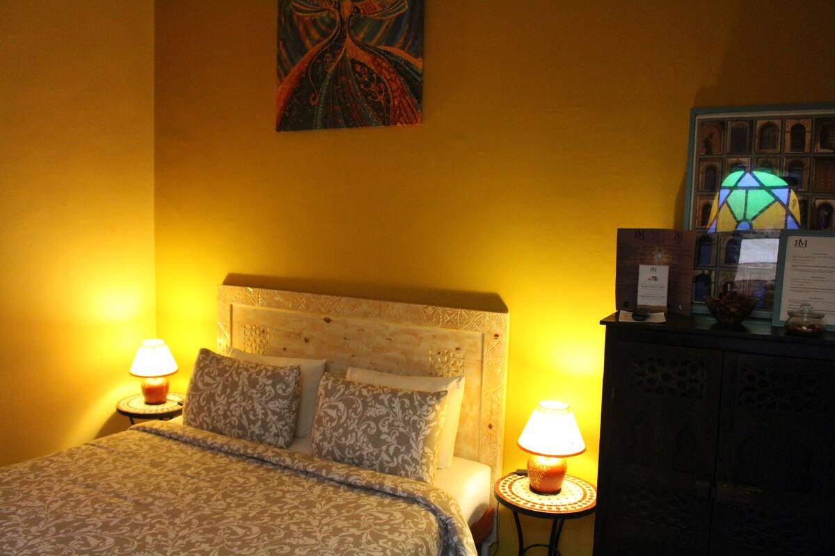 A hotel room with a bed, two bedside lamps, a decorative headboard, a wall painting, and a stained glass window.