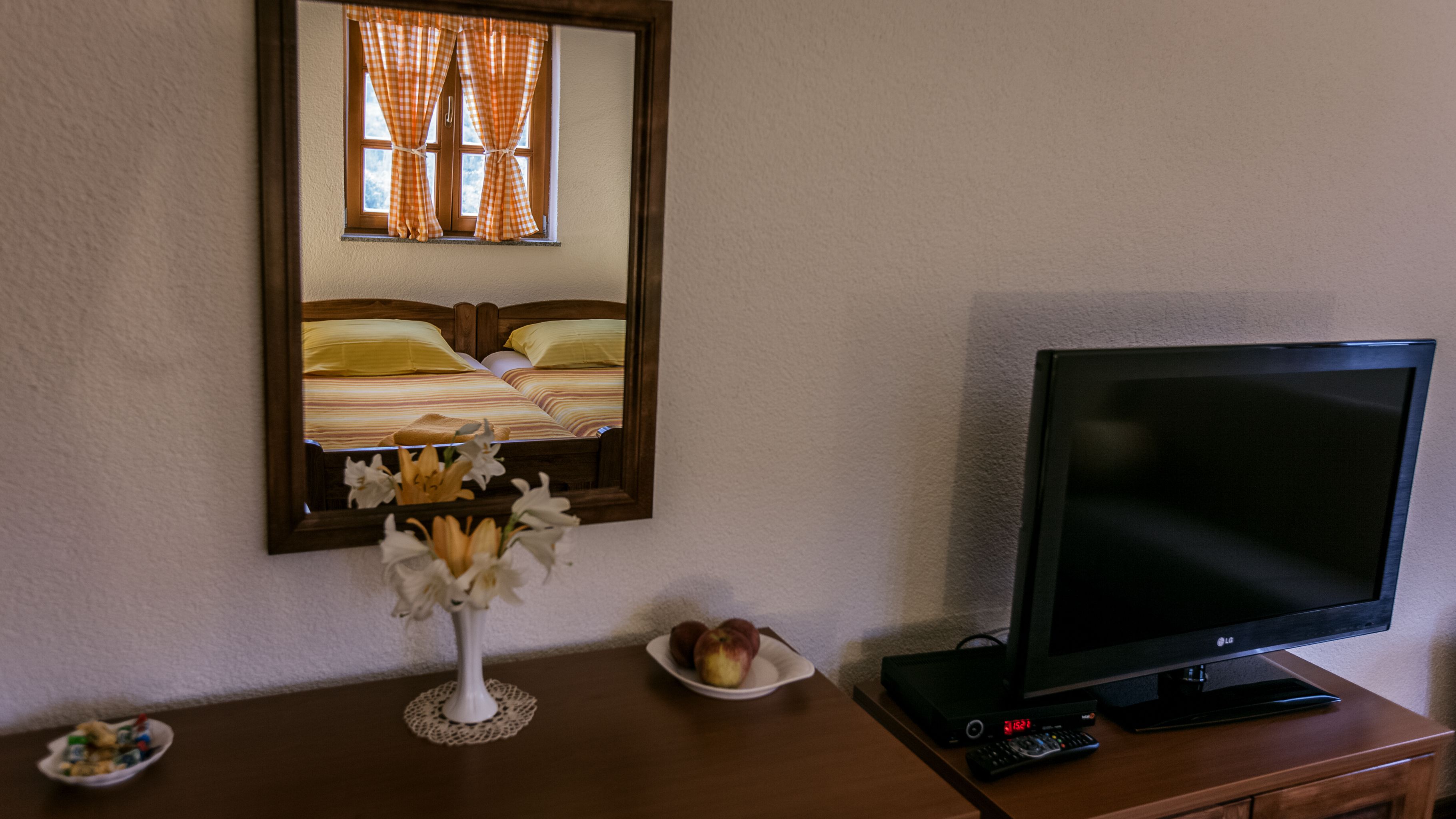 A hotel room with a bed, a television, a vase with flowers, and a plate with fruit.