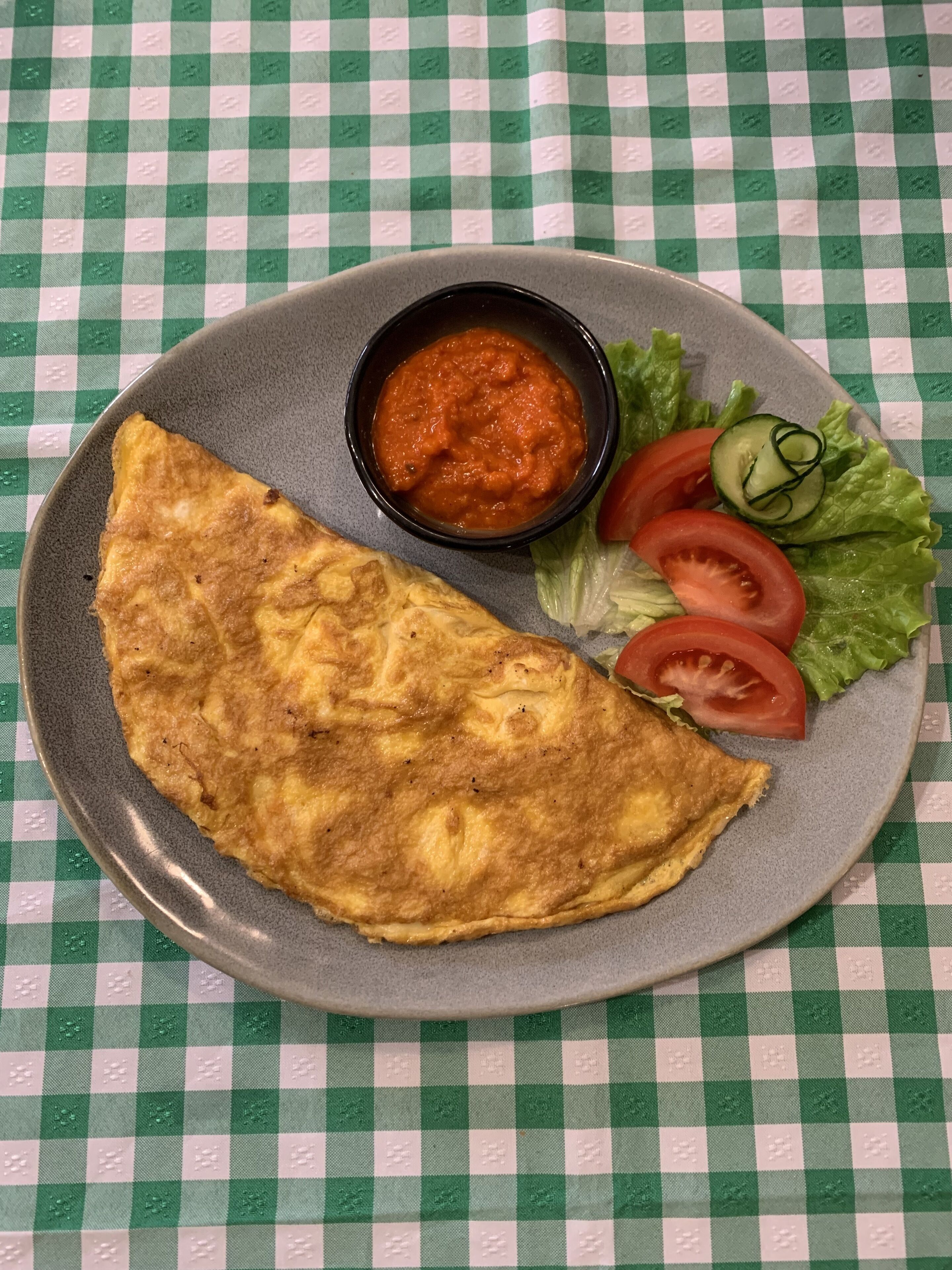 A plate with an omelette, a small bowl of sauce, and fresh vegetables on a checkered tablecloth.