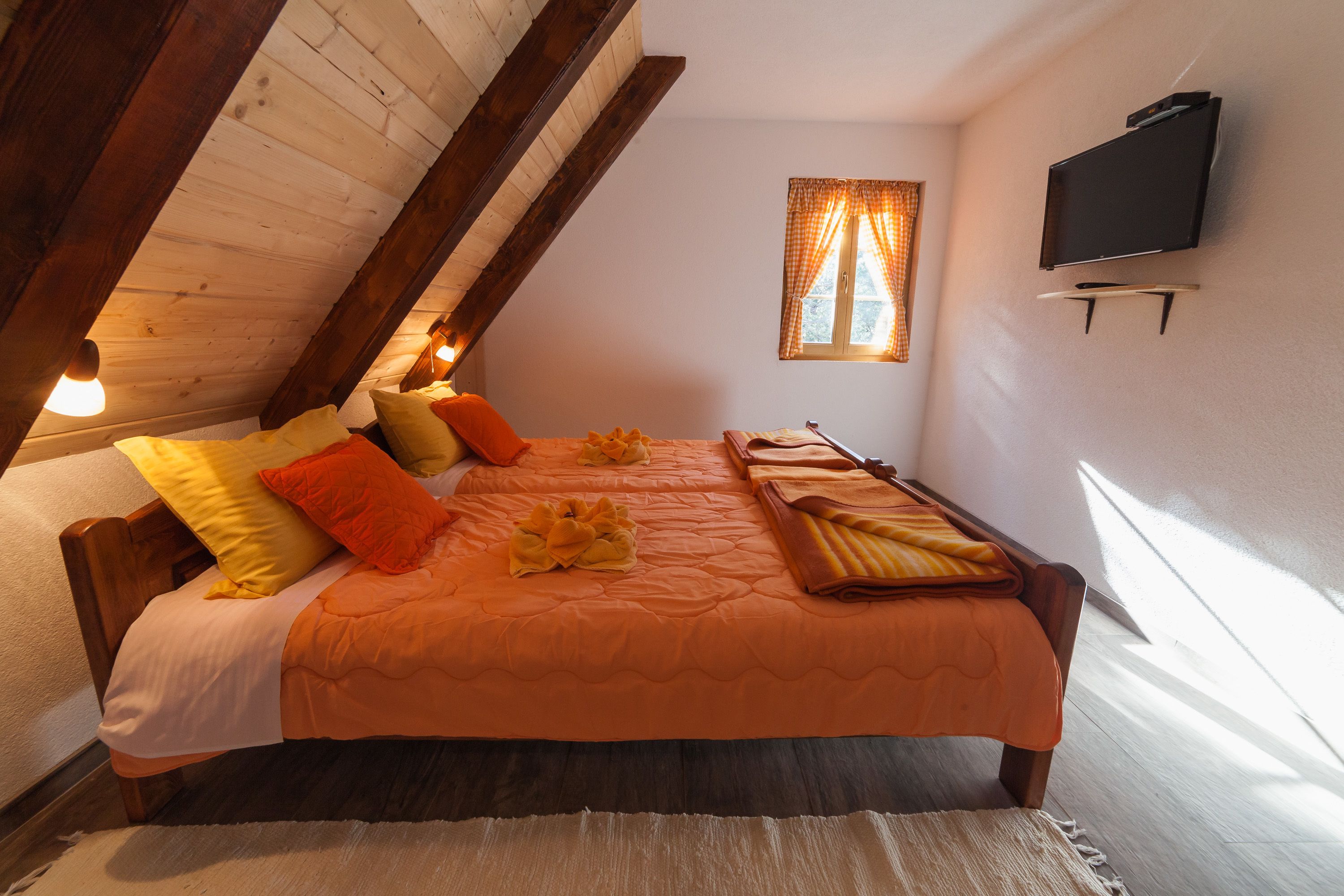 A bedroom with a wooden ceiling, a bed with orange bedding, a television on the wall, and a window with curtains.