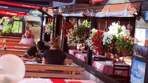 An outdoor café with red and white checkered tablecloths, wooden benches, and hanging flower baskets.