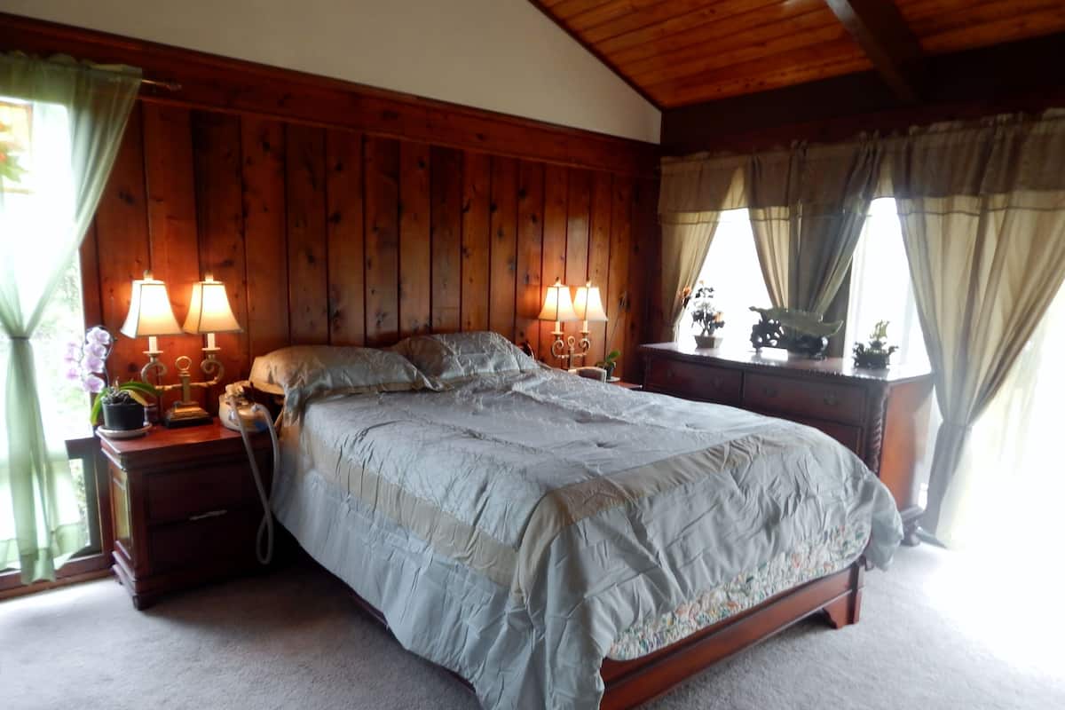 Un dormitorio con cabecera de madera, una cama grande con colcha, dos lámparas de noche, una mesita de noche y una ventana con cortinas.