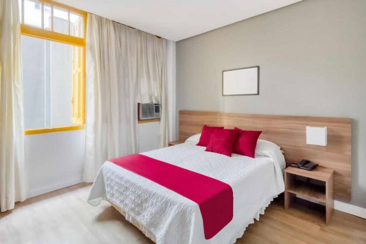 A hotel room with a bed, a bedside table, a nightstand, a window with curtains, and a wall-mounted air conditioning unit.