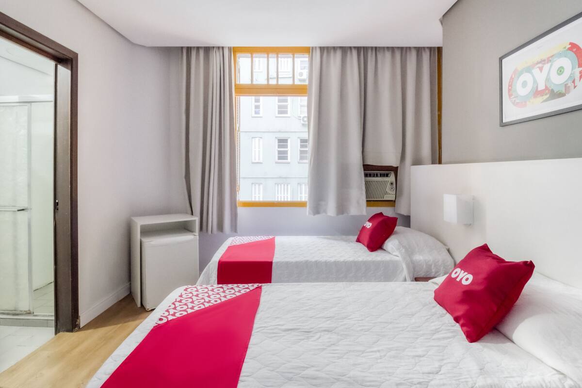 A hotel room with two beds, a window with curtains, a radiator, and a wall-mounted artwork.