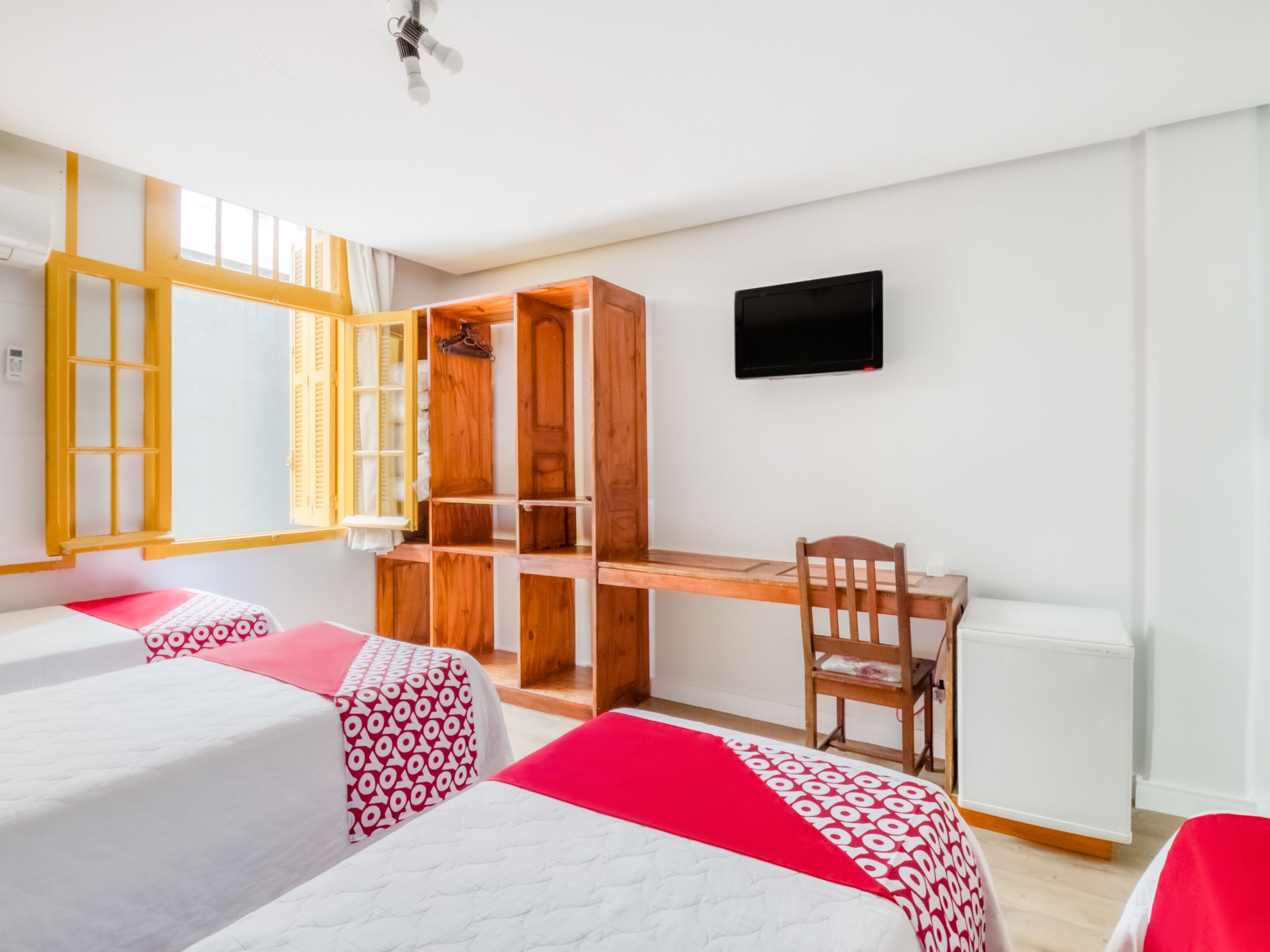 A room with two beds, a wooden desk, a chair, a television, and a window with open shutters.