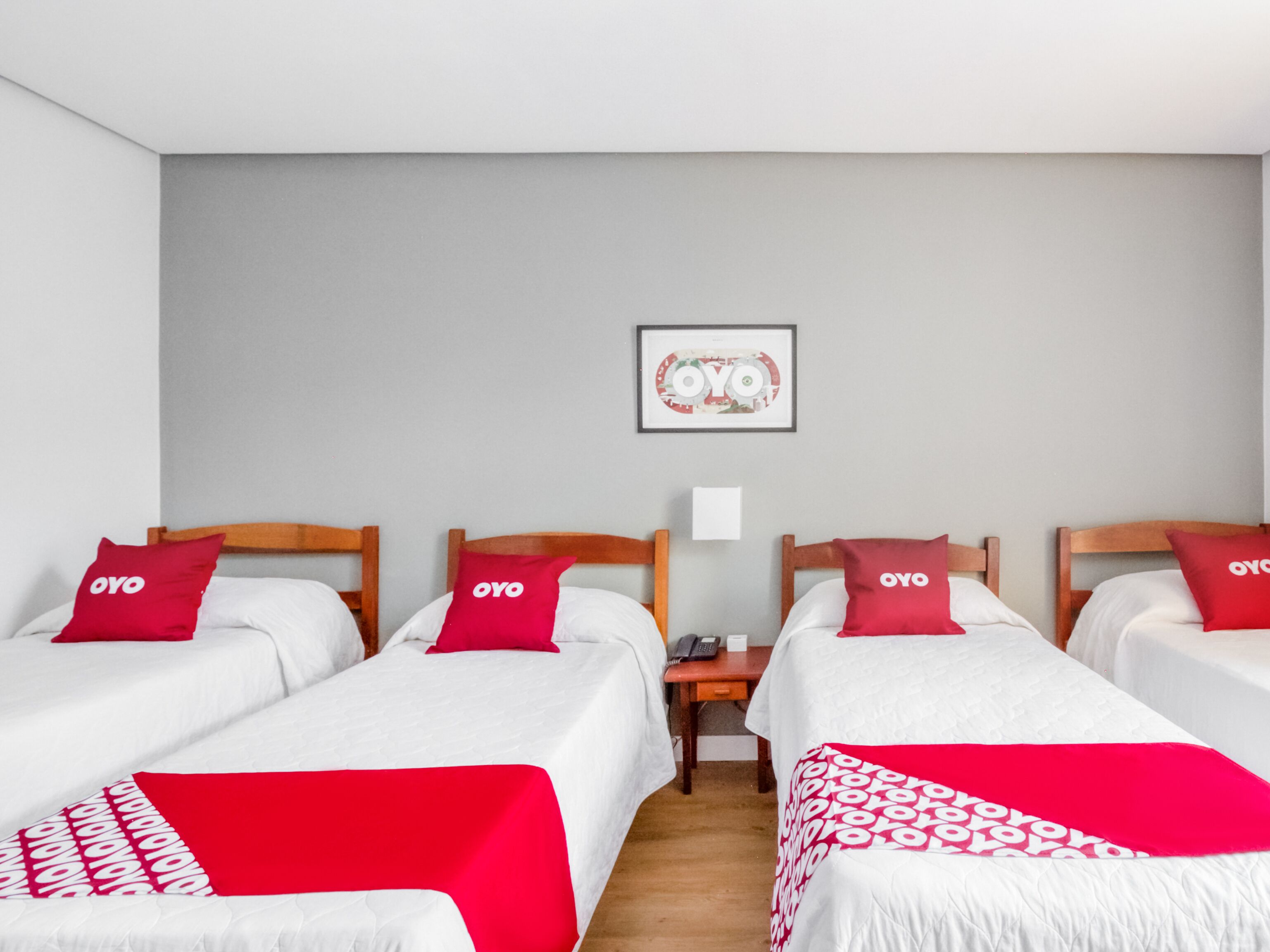 Habitación de hotel con tres camas individuales, cada una con un almohadón rojo y una colcha blanca. Hay una mesita con un teléfono y un cuadro en la pared.