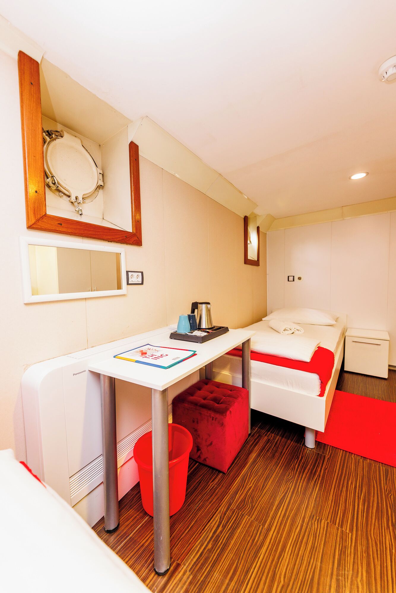 A hotel room with a white bed, a red accent wall, a desk with a coffee maker, and a red trash bin.