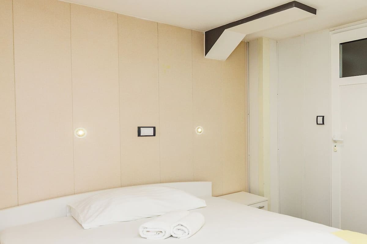 A neatly made bed with white linens, a yellow blanket, and a folded towel. There are recessed ceiling lights and a small picture frame on the wall.