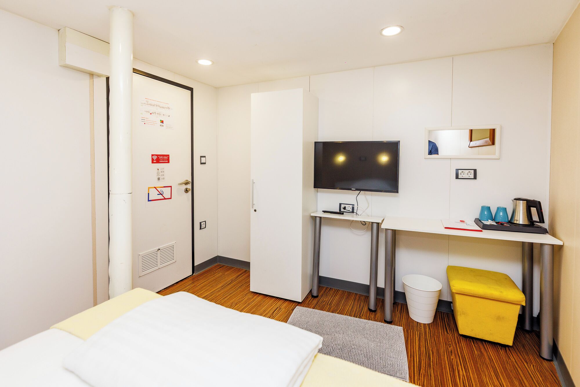 Habitación de hotel con cama, escritorio con televisor, un pequeño cubo de basura y un reposapiés amarillo.