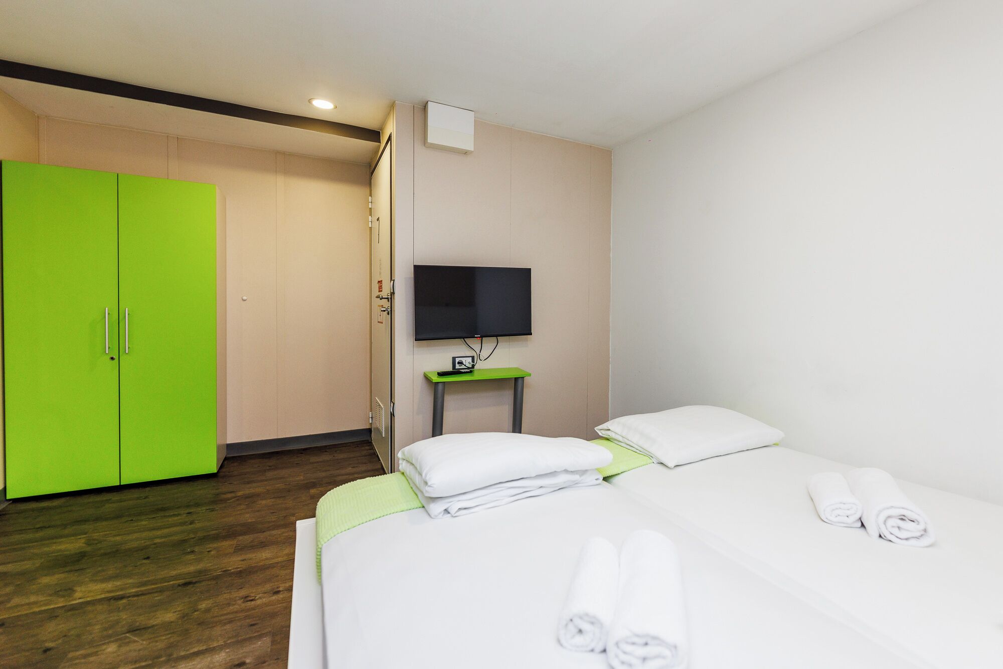 Une chambre d’hôtel avec un lit, une armoire verte, un téléviseur posé sur un meuble et une petite table.