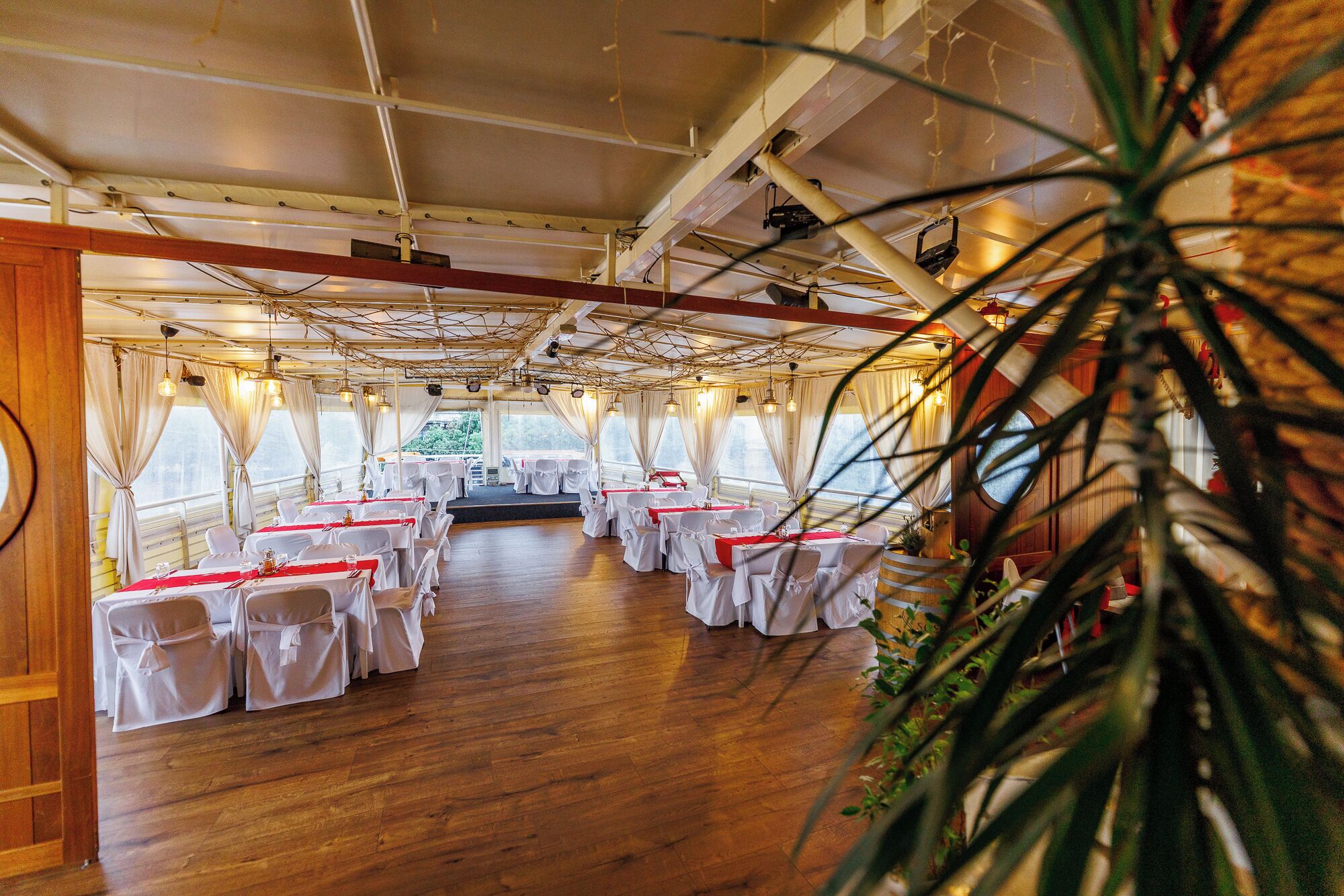 Un comedor con mesas dispuestas para un evento formal, con manteles blancos y sillas, suelo de madera y ventanas grandes con cortinas.
