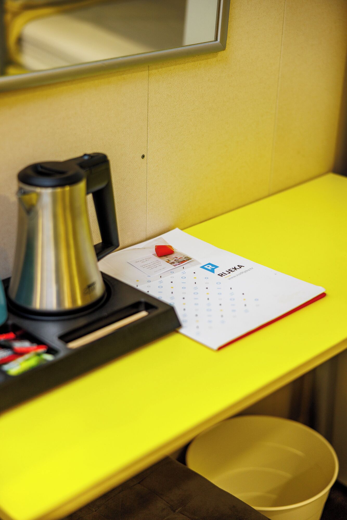 Un escritorio amarillo con una cafetera, una libreta roja y un cubo de basura amarillo.