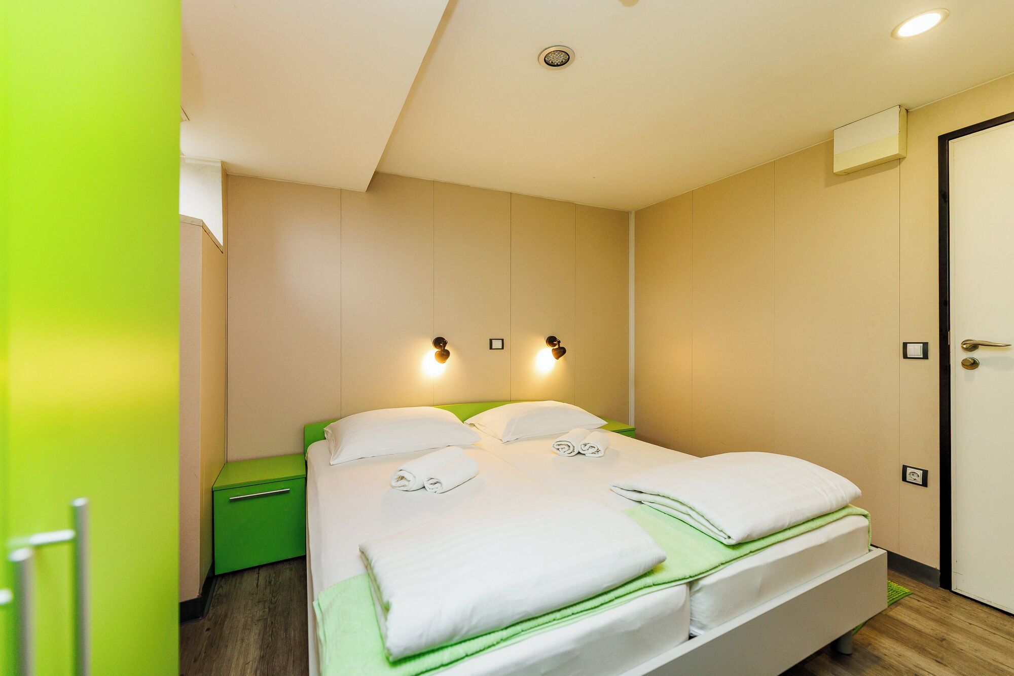 Habitación de hotel con una cama, mesitas de noche verdes, una puerta y luces fijadas a la pared.