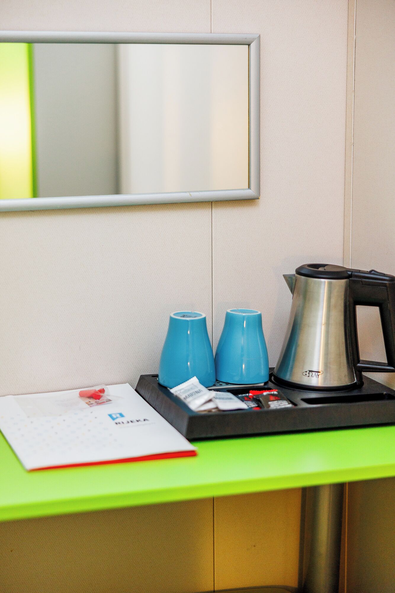 Habitación de hotel con un escritorio verde, un espejo, dos tazas azules, un hervidor plateado y un menú en la encimera.