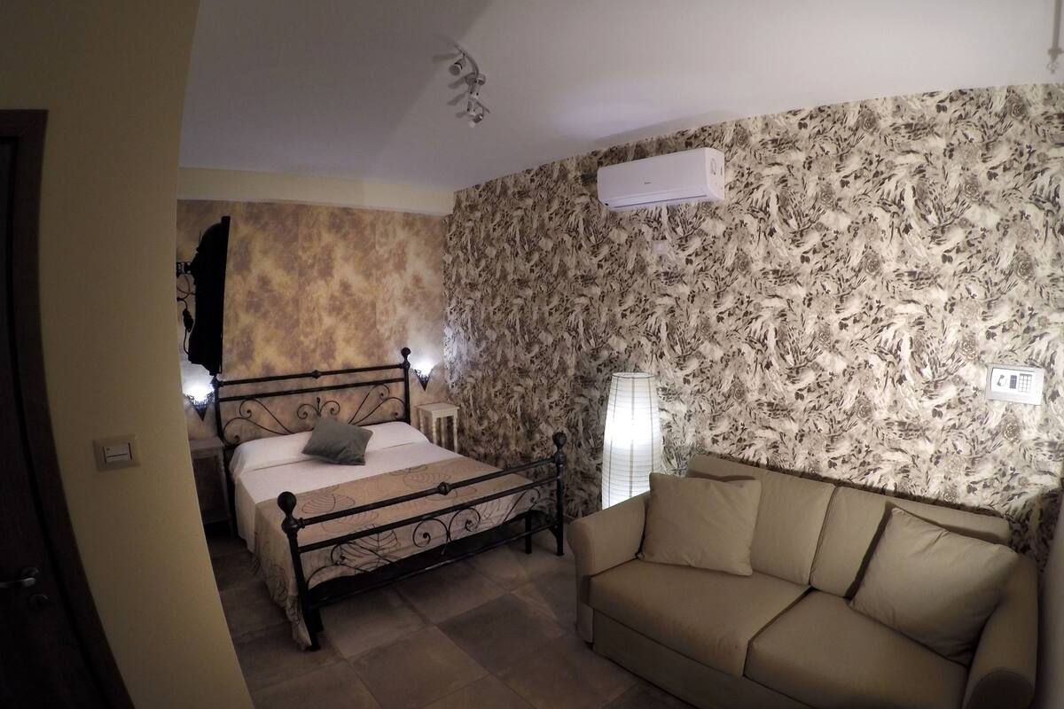 A bedroom with a bed, a bedside table, a lamp, a sofa, and a wall-mounted air conditioner.
