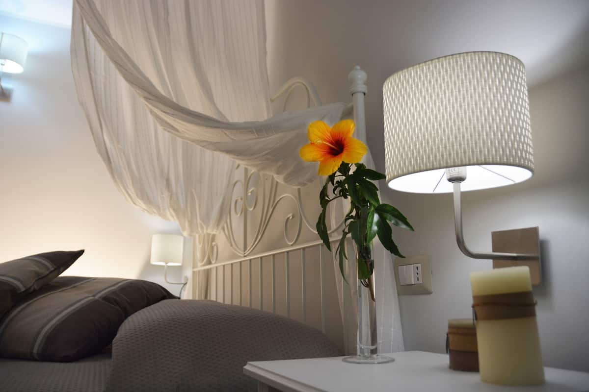 A bedroom with a bed, a bedside table with a lamp, a vase with a flower, and a wall-mounted switch.