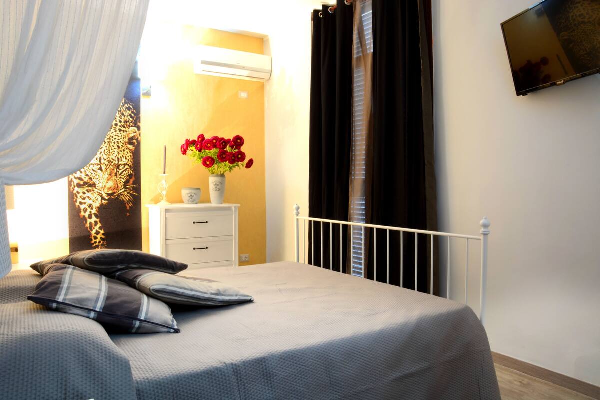 A bedroom with a bed, pillows, a nightstand, a vase with flowers, a wall-mounted TV, and a window with curtains.