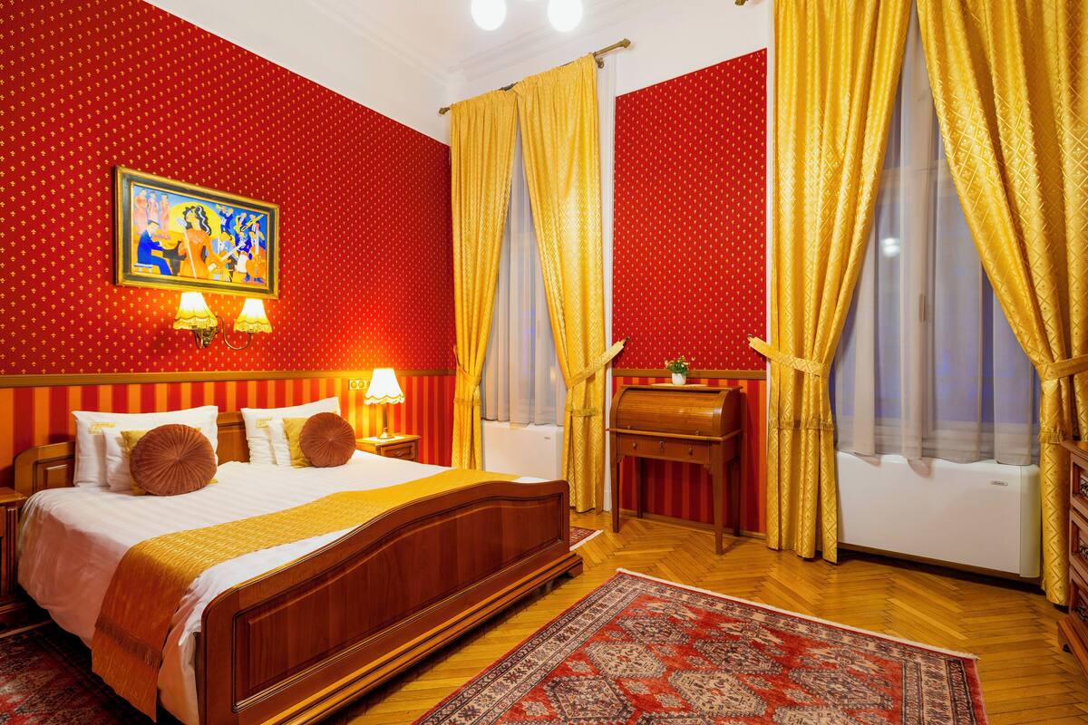 Una habitación de hotel con una cama grande, una mesita de noche de madera con lámparas, una pared con estampado rojo y un cuadro.