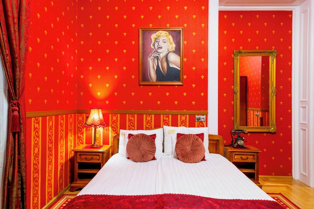 Habitación de hotel con pared de diseño rojo y dorado, cama con ropa de cama blanca, dos almohadas marrones, mesita de noche con lámpara y espejo en la pared.