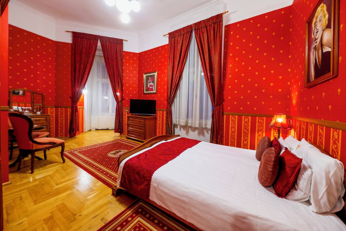 Habitación de hotel con piso de madera, una cama con ropa de cama blanca y roja, un escritorio con silla, un televisor y un cuadro en la pared.