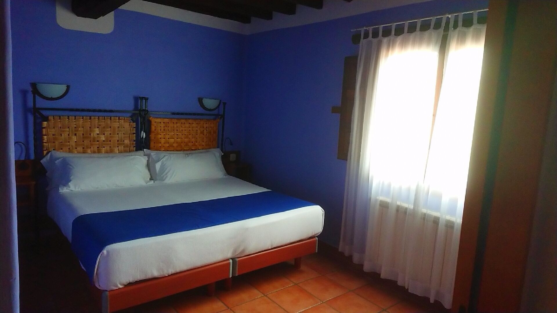 Un dormitorio con cama, cabecera, mesita de noche, lámpara y ventana con cortinas.