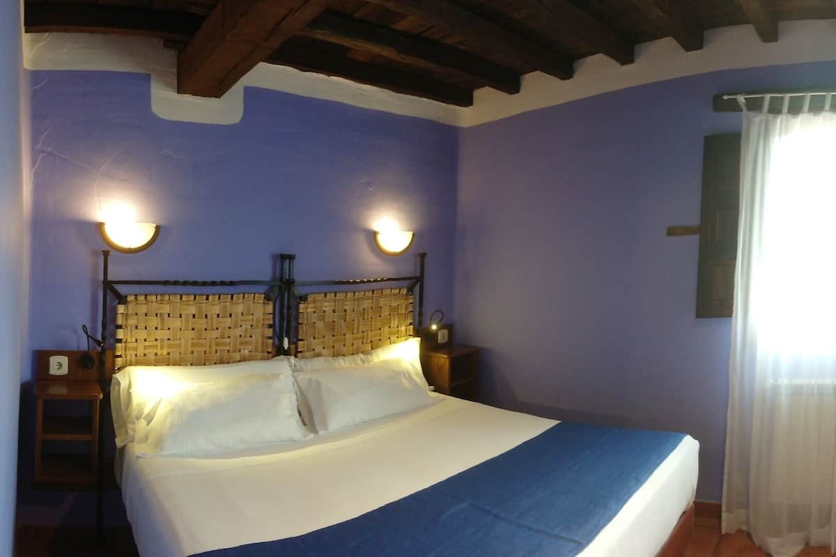 Quarto com cama, duas luminárias fixadas na parede, janela com cortinas leves, cadeira de madeira e uma mesa pequena.