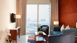 Een hotelkamer met een groot raam, een flatscreen televisie, een bank, een stoel, een salontafel en een vaas met bloemen.