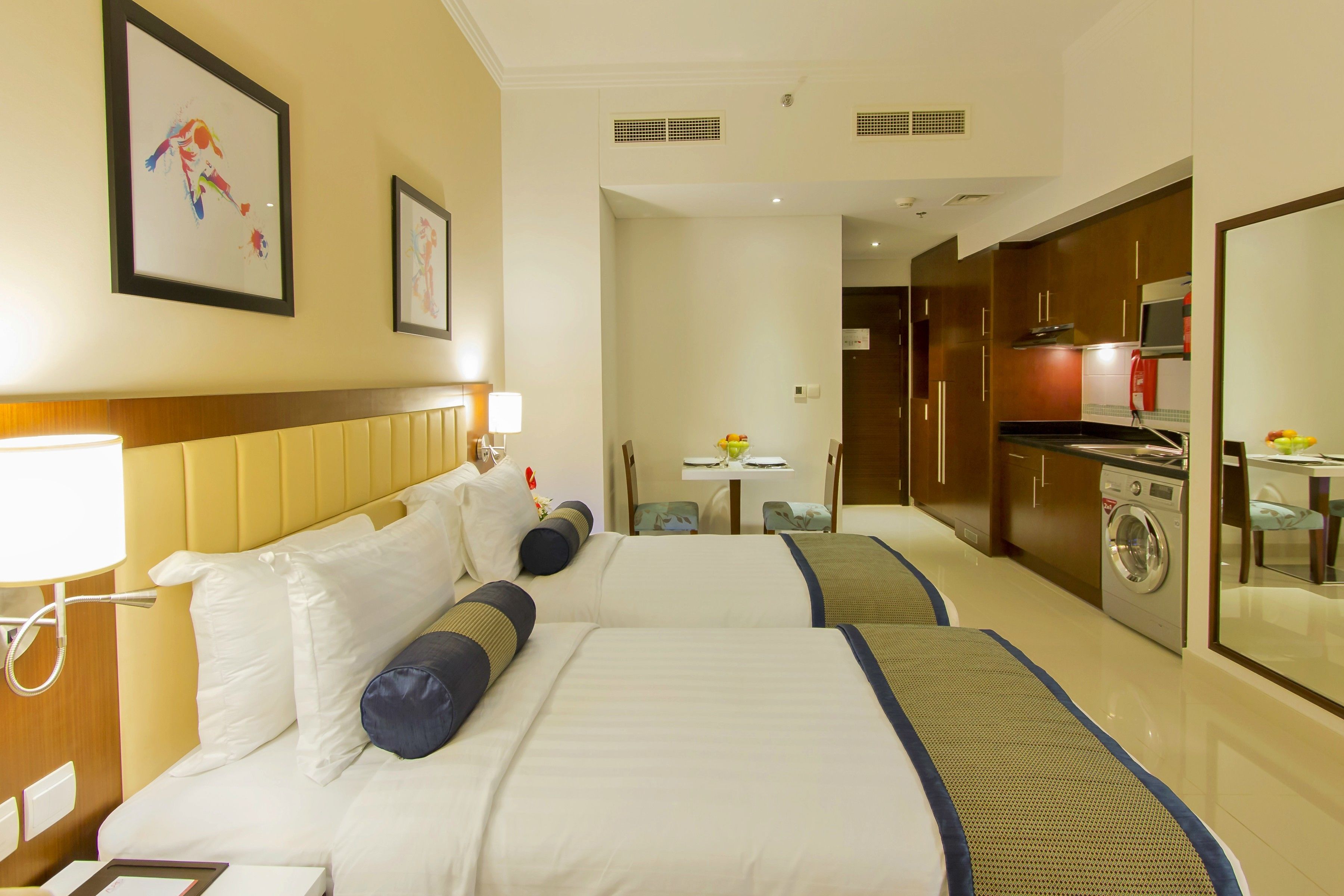 A hotel room with two beds, a small kitchenette, a dining area, and a bathroom.