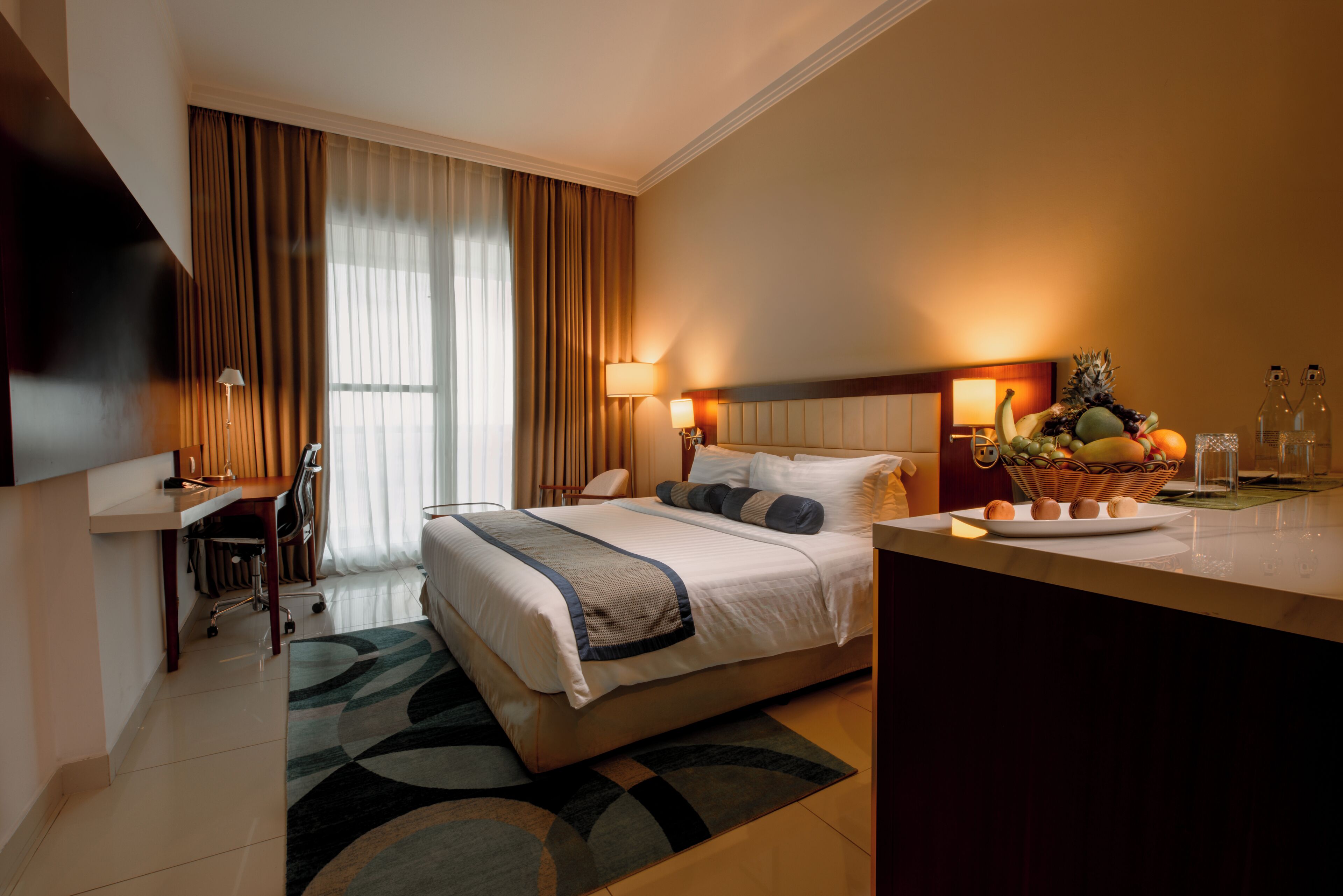 Een hotelkamer met een bed, bureau, stoel en een fruitmandje op het nachtkastje.