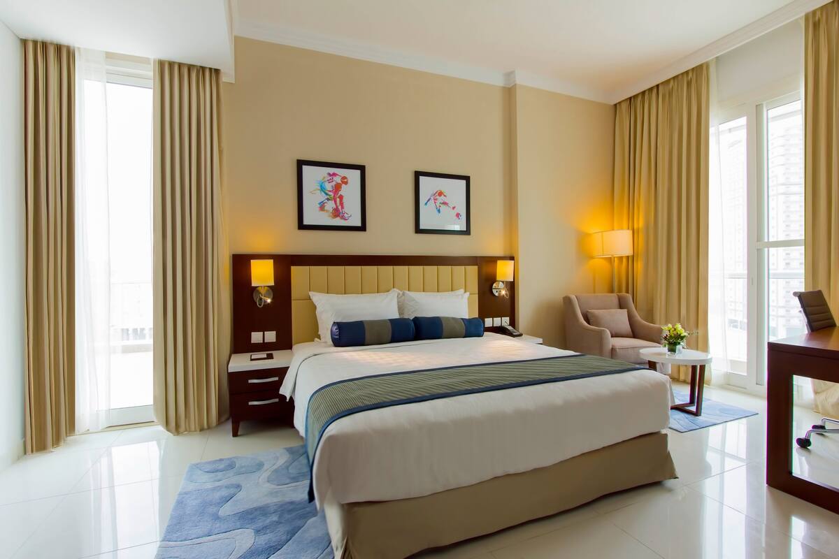 A hotel room with a large bed, a desk, a chair, and two framed artworks on the wall.