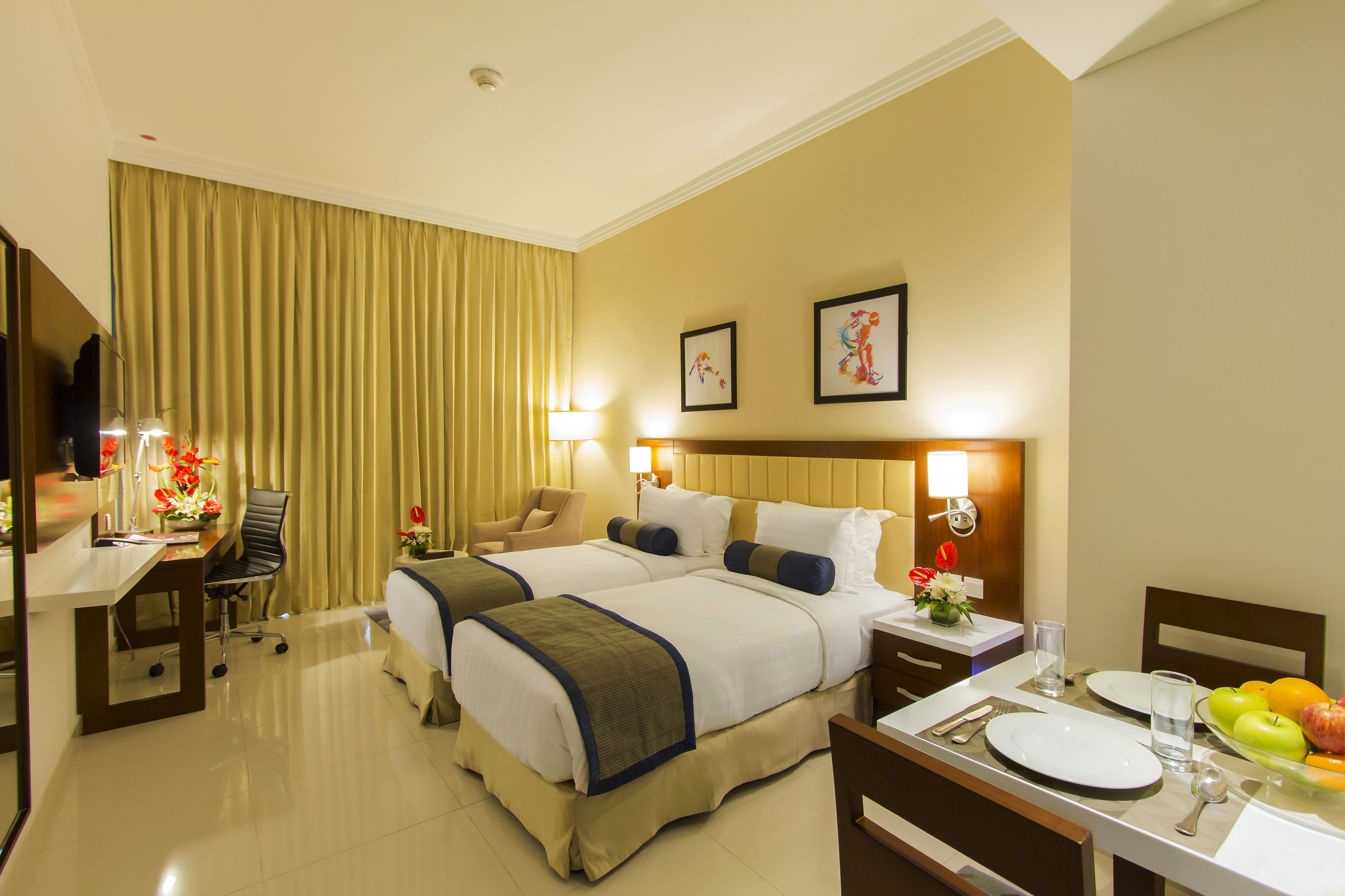 A hotel room with two beds, a desk with a chair, a dining table set for two, and a TV.