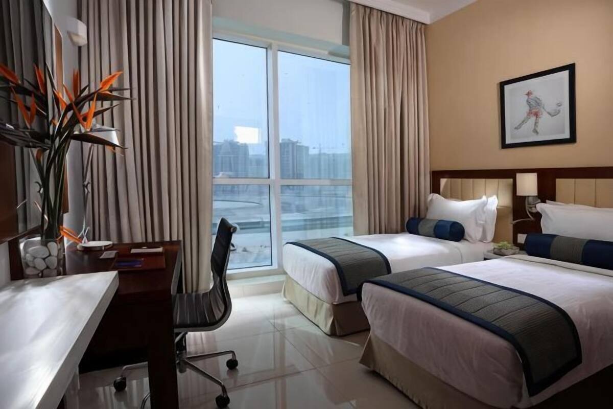 A hotel room with two beds, a desk, a chair, a large window with curtains, and a framed picture on the wall.