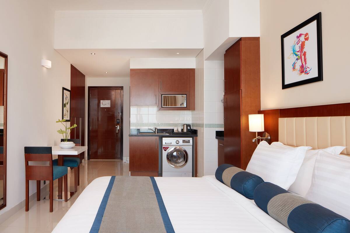 A hotel room with a bed, a desk with a chair, a washing machine, and a kitchenette.