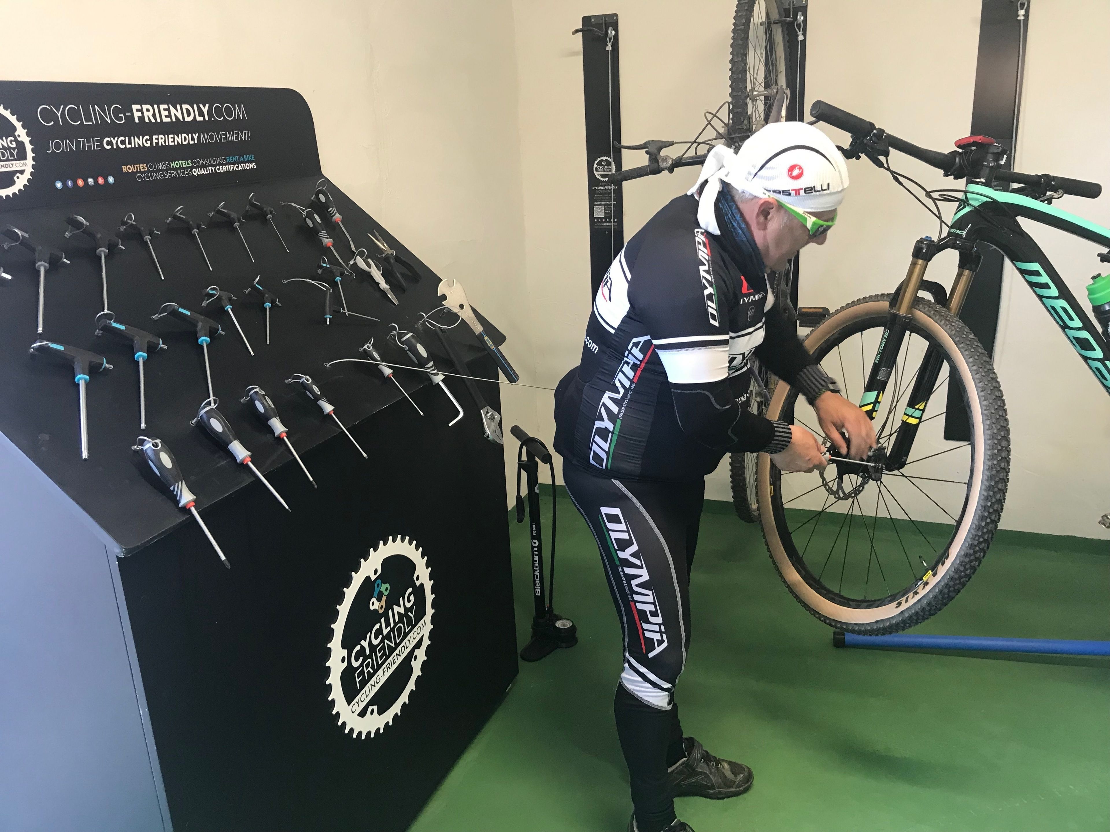A cyclist in cycling gear working on a bike in a room with a wall-mounted tool display and a bike rack.