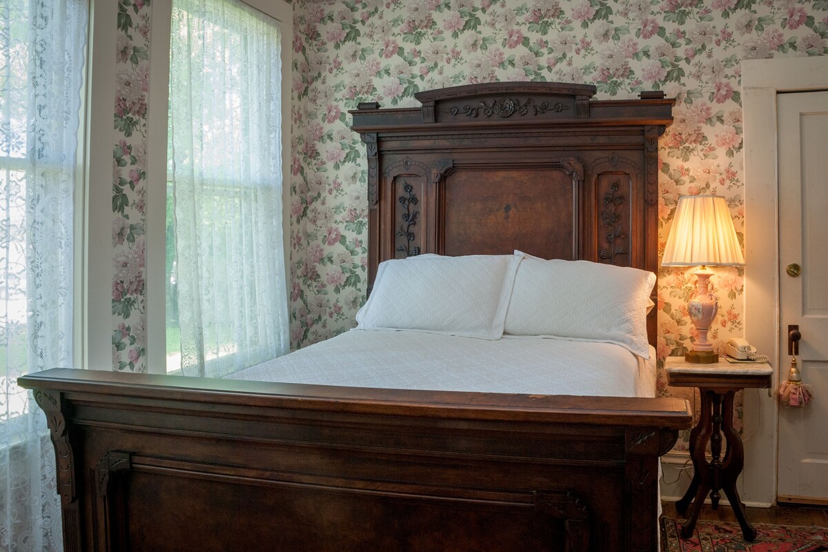 A traditional wooden bed with a headboard, a bedside table with a lamp, and floral wallpaper.