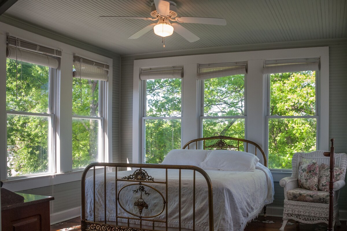 A bedroom with a wooden bed, a chair, and large windows with a view of trees.