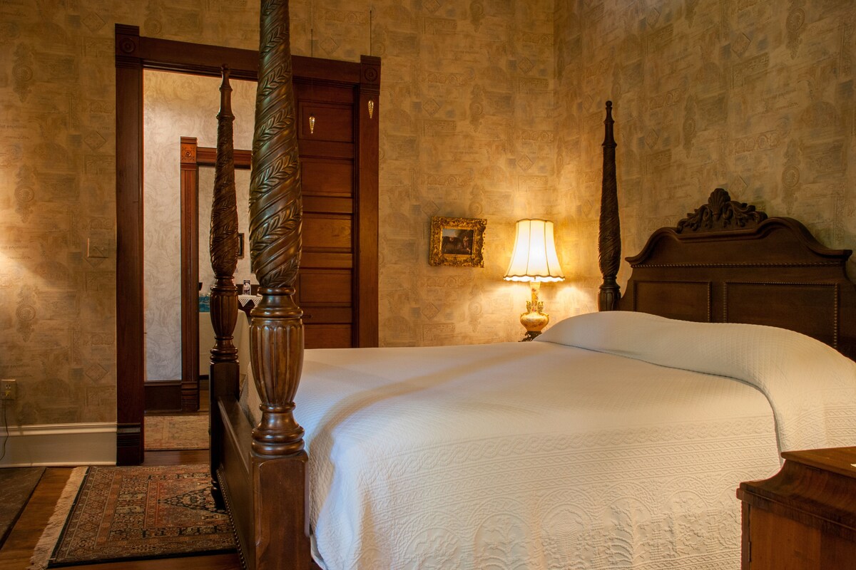 A traditional bedroom with a four-poster bed, a wooden headboard, a bedside lamp, and a patterned rug.