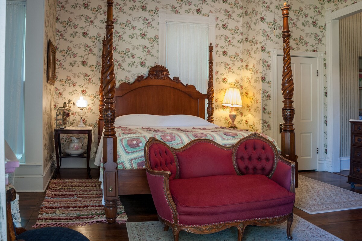A bedroom with a four-poster bed, a red upholstered chair, and floral wallpaper.