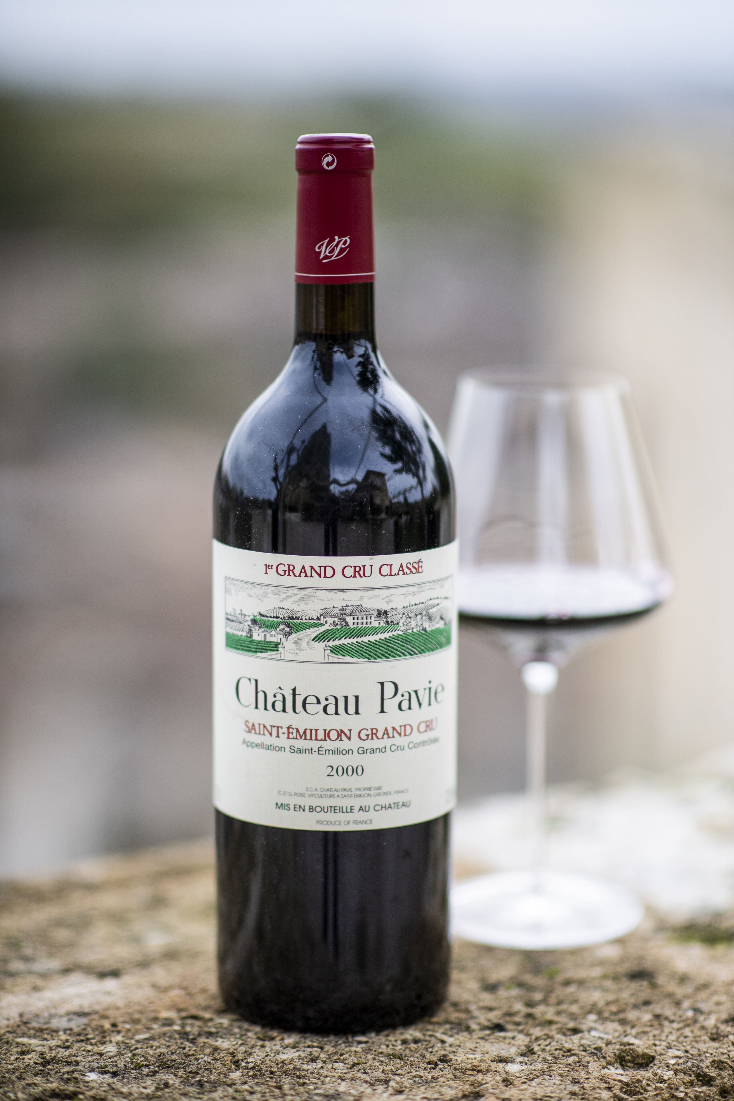 Una botella de vino Château Pavie con etiqueta, sobre una superficie de piedra con una copa de vino al lado.