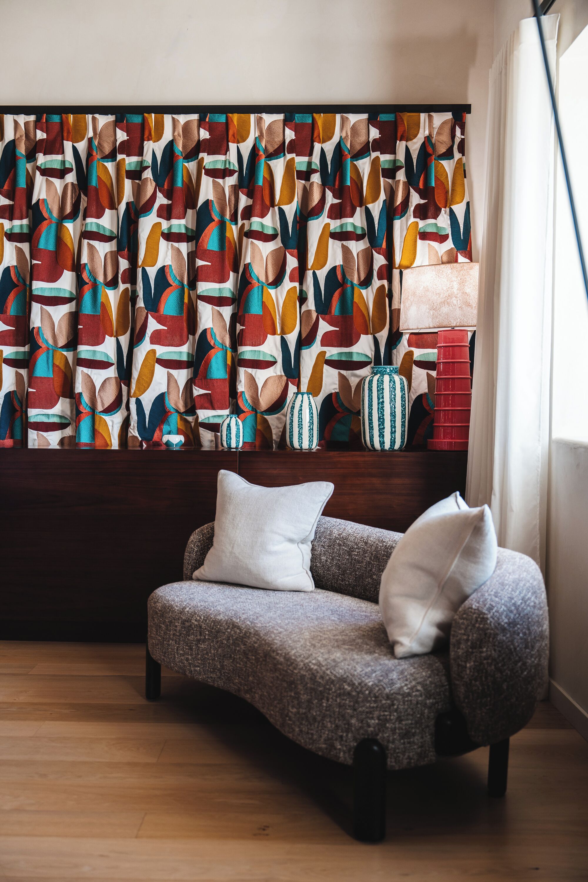 A cozy living room with a patterned curtain, a gray sofa with cushions, and decorative vases on a wooden shelf.