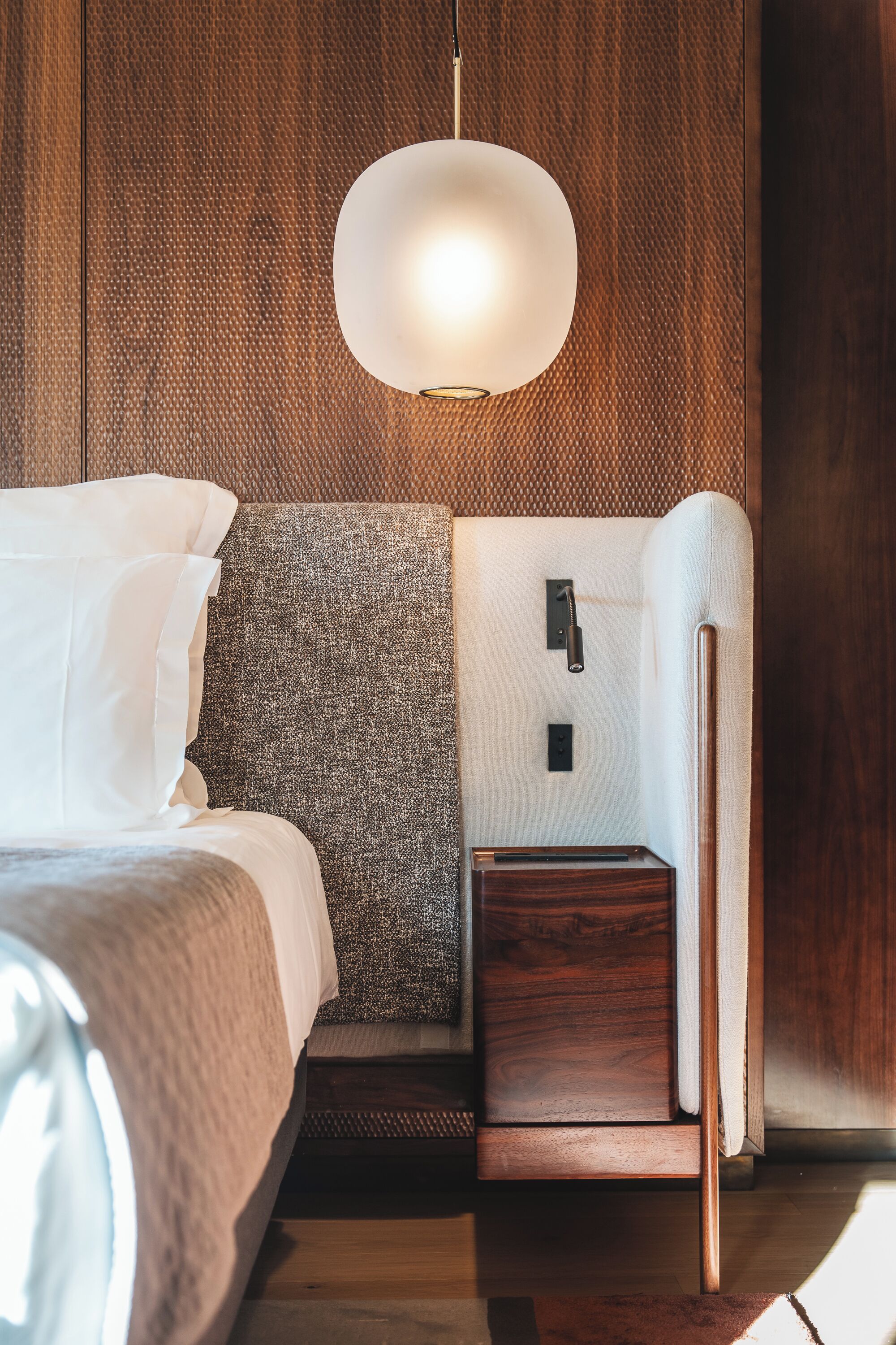 A modern hotel room with a wooden wall, a round light fixture, a bed with white linens, a grey upholstered bench, and a small wooden nightstand.