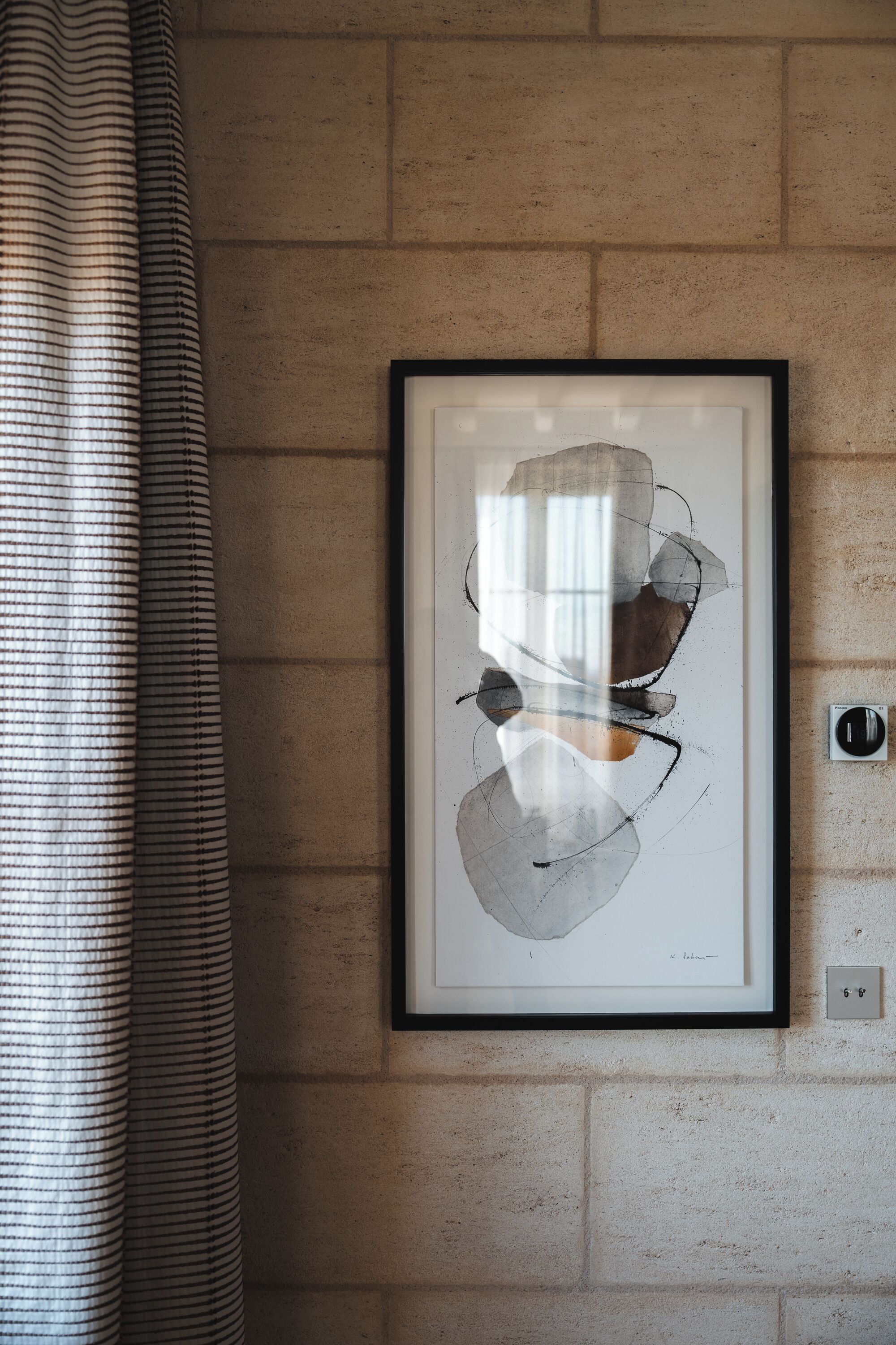 A framed abstract artwork on a brick wall, a power outlet, and a striped curtain.