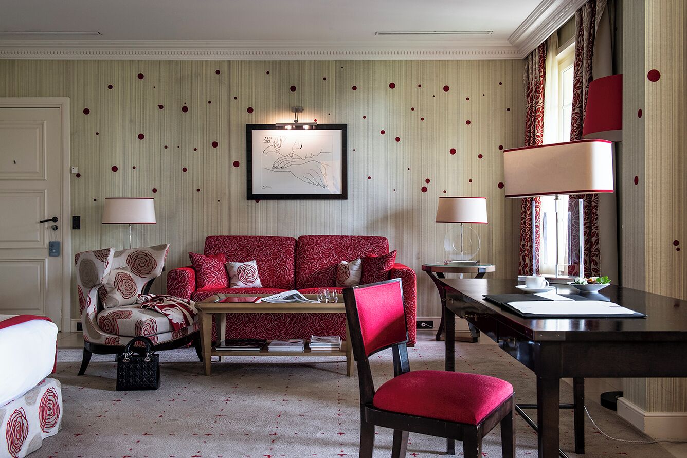 A living room with a red sofa, a coffee table, a chair, and a dining table. There is a framed artwork on the wall and a window with curtains.