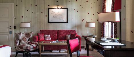 A living room with a red sofa, a coffee table, a chair, and a dining table. There is a framed artwork on the wall and a window with curtains.