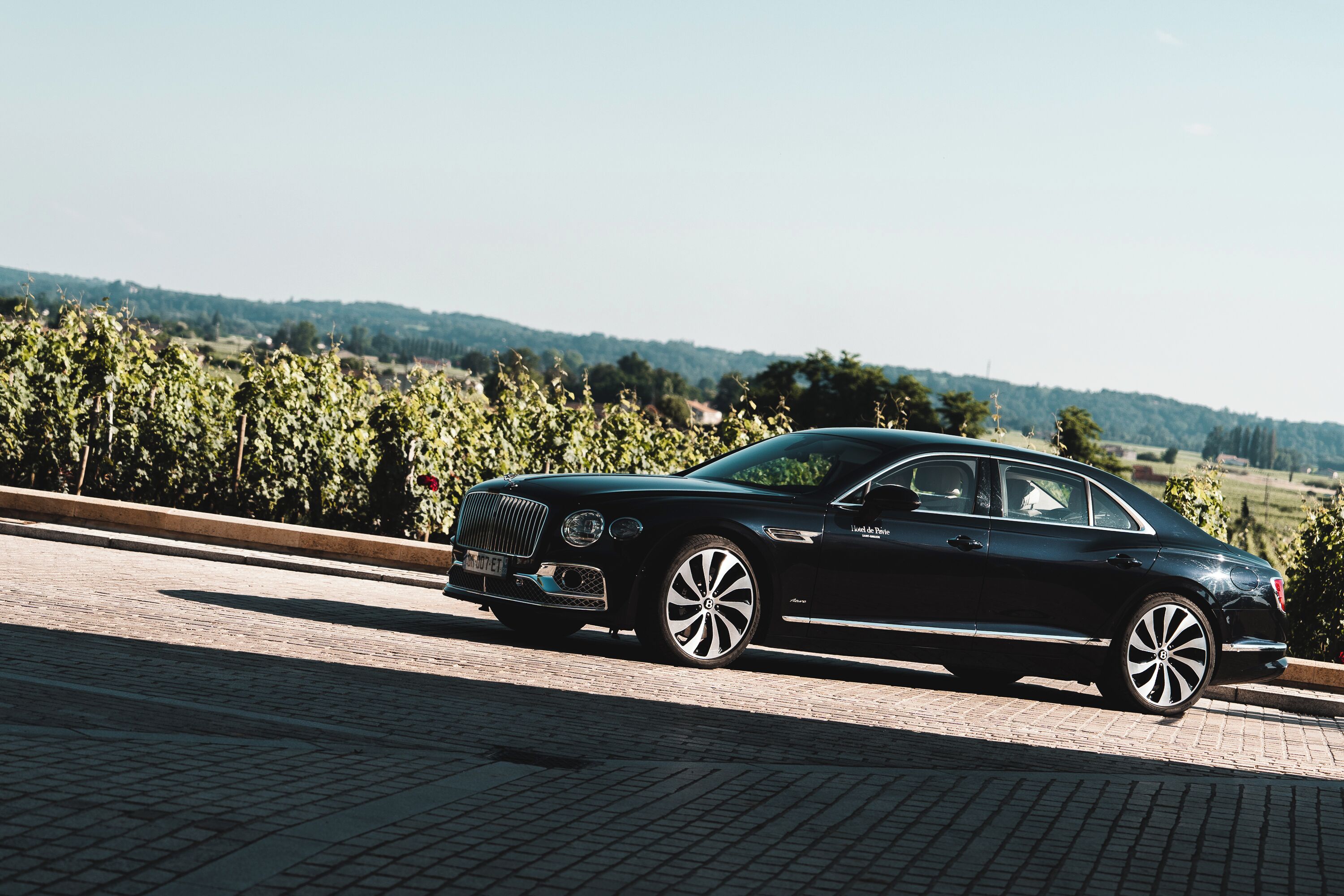 A black luxury car parked on a paved road with a vineyard in the background.